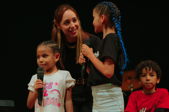 niñas cantando en una tarima y profesora dándoles apoyo emocional