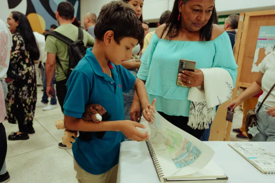 Niño estudiante de Musicarte revisando con orgullo su bitácora de dibujos en una exposición, ilustrando el desarrollo del pensamiento crítico y la creatividad a través de las artes visuales.