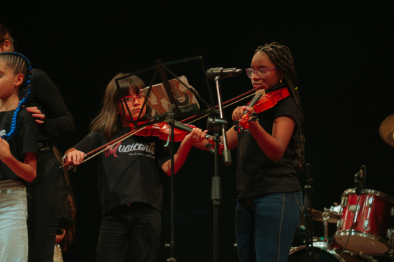 Estudiantes de Musicarte tocando el violín concentradas en un escenario, demostrando los beneficios de la educación musical para el desarrollo de la disciplina y la confianza en niños.